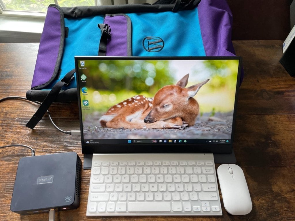 My mini pc setup with a portable monitor, small keyboard and mouse, and a purple and teal backpack behind it to carry it all in.